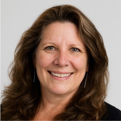 Beth S. Rous smiling, with long brown wavy hair, in front of a light gray background